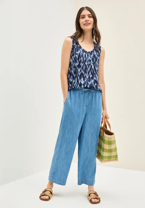 Frau mit gemustertem Top, blauer Hose und Tasche, trägt Sandalen.